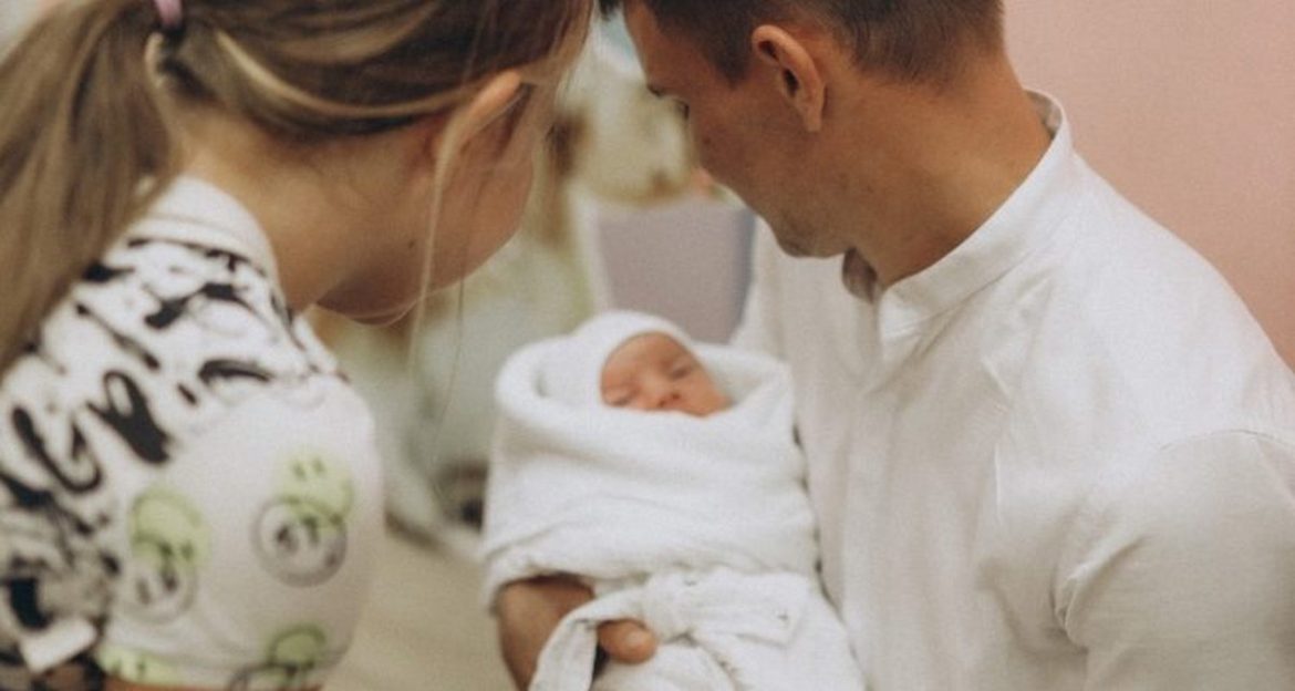 Рождаемость в Беларуси упала почти в два раза – тревожная статистика в мировом контексте Мужчина держит младенца на руках, женщина склонилась рядом – семья после роддома.