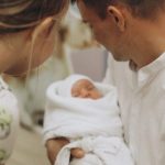 Мужчина держит младенца на руках, женщина склонилась рядом – семья после роддома.