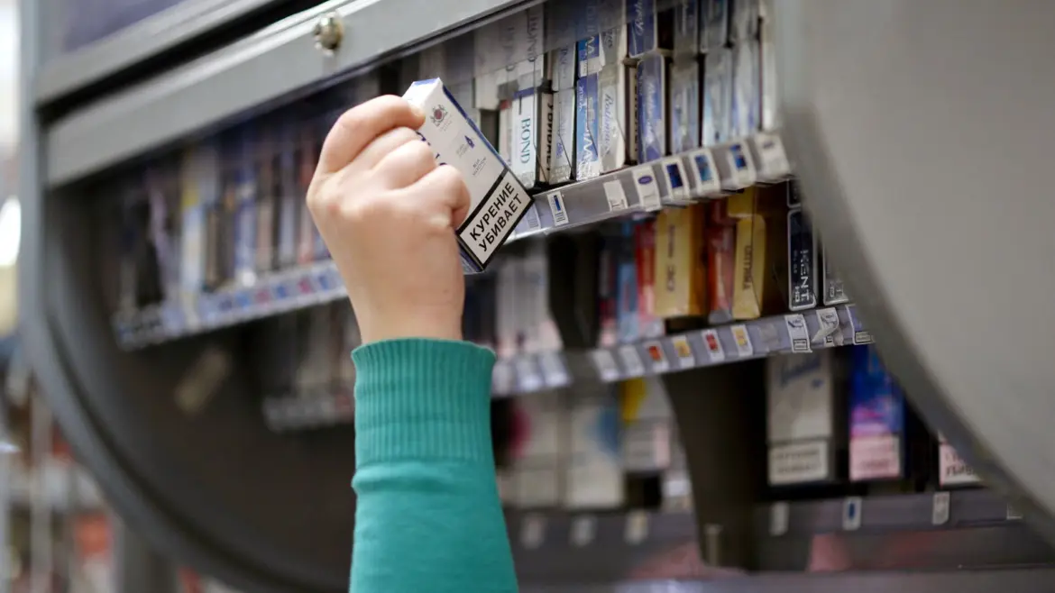 Рука покупателя тянется к пачке сигарет на витрине в магазине.