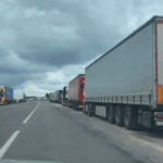 Длинная колонна грузовиков многополосной дороге, небо затянуто серыми тучами.