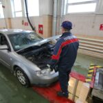 Сотрудник в форме "Гостехосмотра" осматривает автомобиль на станции технического осмотра – капот открыт, машина стоит у смотровой ямы в помещении.