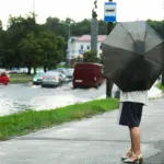 Женщина с чёрным зонтам стоит на тротуаре в дождливый день в Минске, машины едут по затопленной дороге, на горизонте – городской пейзаж