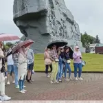 Группа туристов с экскурсоводом у мемориала «Брестская крепость», экскурсия проходит под дождём, люди с зонтами слушают рассказ гида