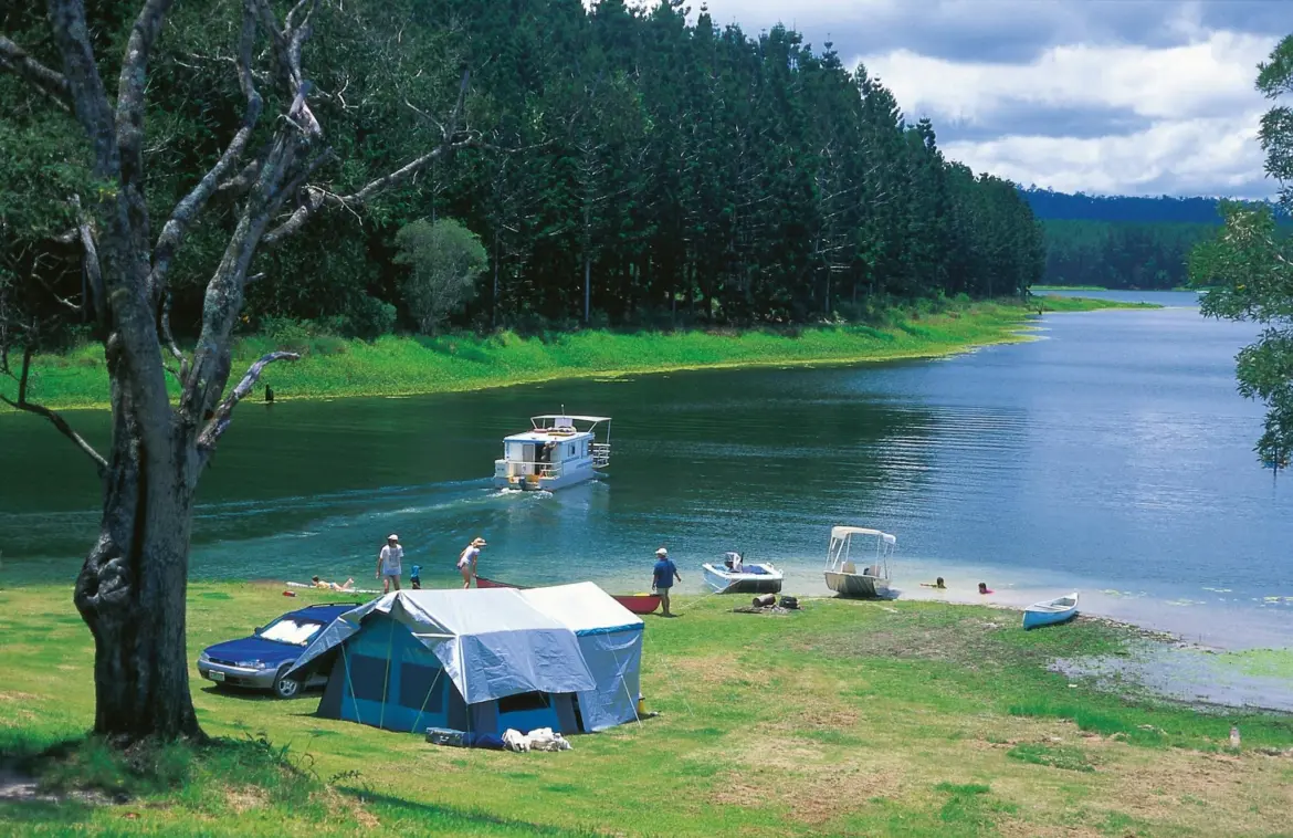 Люди отдыхают у палаточного лагеря на берегу реки, рядом лодки и катер, зелёный лес на фоне, прогноз обещает дожди и прохладу