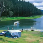 Люди отдыхают у палаточного лагеря на берегу реки, рядом лодки и катер, зелёный лес на фоне, прогноз обещает дожди и прохладу