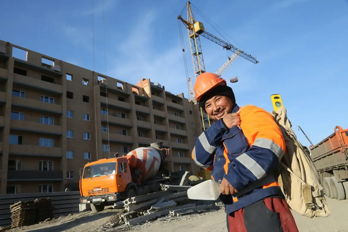 Кадровый голод по-белорусски: импортируем рабочих из Пакистана Рабочий в оранжевом жилете и каске на строительной площадке в Беларуси на фоне дома и строительной техники.