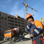 Рабочий в оранжевом жилете и каске на строительной площадке в Беларуси на фоне дома и строительной техники.