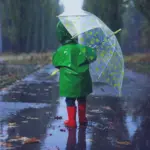 Ребёнок в зеленом дождевике с зонтом бежит по мокрой дороге