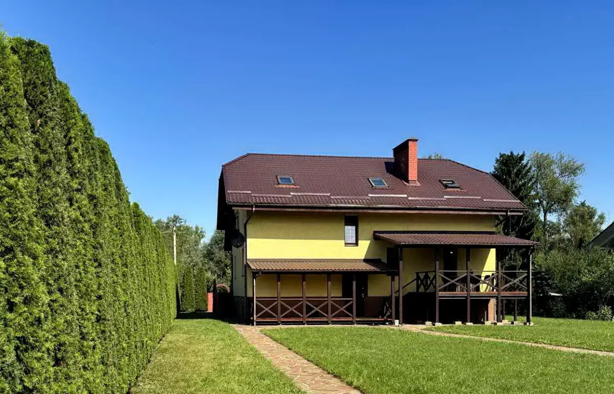 Дом с мансардой и верандой на зелёном участке в Клейниках, рядом с живой изгородью из туй, на фоне чистого неба.