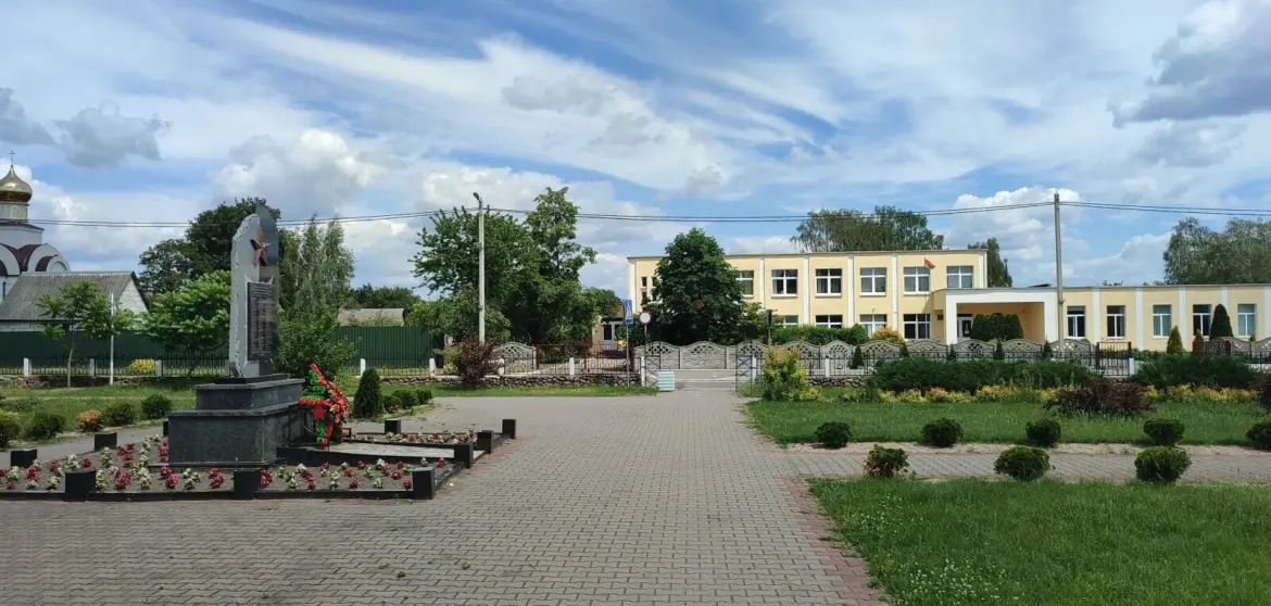 Площадка в центре Клейников с памятником, школой и благоустроенной территорией, на фоне зелени и голубого неба.