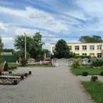 Площадка в центре Клейников с памятником, школой и благоустроенной территорией, на фоне зелени и голубого неба.