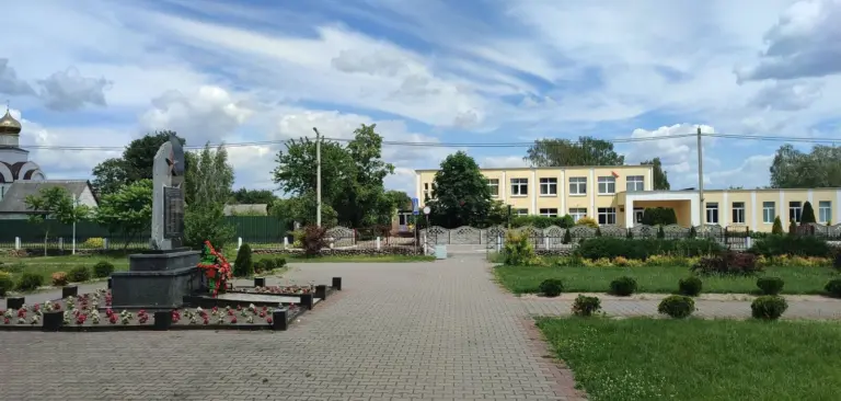 Площадка в центре Клейников с памятником, школой и благоустроенной территорией, на фоне зелени и голубого неба.