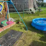 На фото двор частного дома: у стены стоит скамейка с компрессором и удлинителем, рядом расстелен коврик, чуть дальше – надувной бассейн