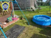На фото двор частного дома: у стены стоит скамейка с компрессором и удлинителем, рядом расстелен коврик, чуть дальше – надувной бассейн