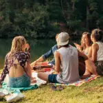 Группа друзей сидит на берегу реки, беседует и слушает музыку на пикнике.