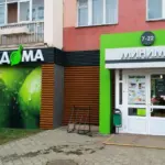 Вход в магазин «У дома» с зелёной вывеской и витриной с изображением зелёных яблок.