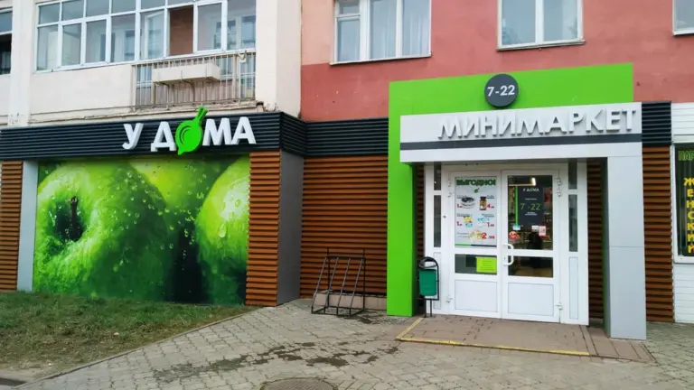 Вход в магазин «У дома» с зелёной вывеской и витриной с изображением зелёных яблок.